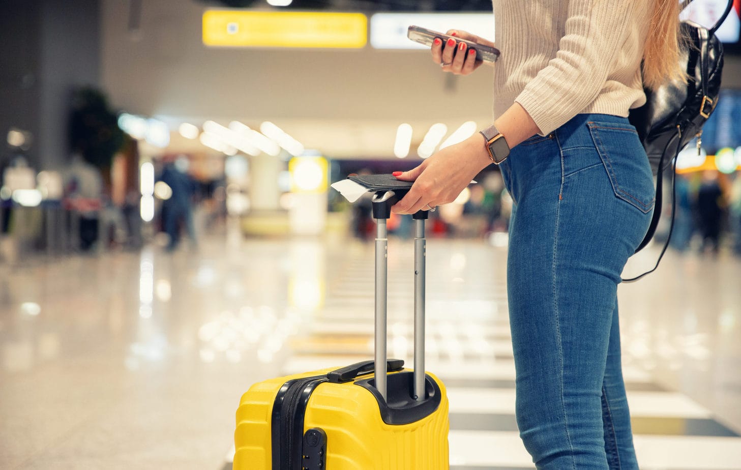 Woman holding a smartphone with a boarding pass at an airport terminal, standing next to a yellow wheeled suitcase.