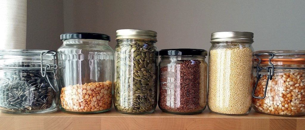A variety of food items stored in clear glass jars with different lids, showcasing sunflower seeds, split peas, pumpkin seeds, flax seeds, millet, and popcorn kernels on a wooden shelf.